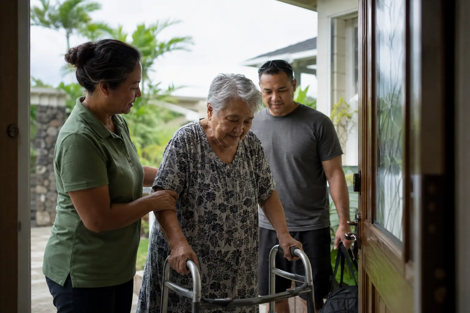 A caregiver steadying an older woman using a walker as she arrives home, her son close behind